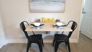 En matplats med ett träbord, två svarta stolar, en skylt med texten ”Hej!” och en målning av en strand.