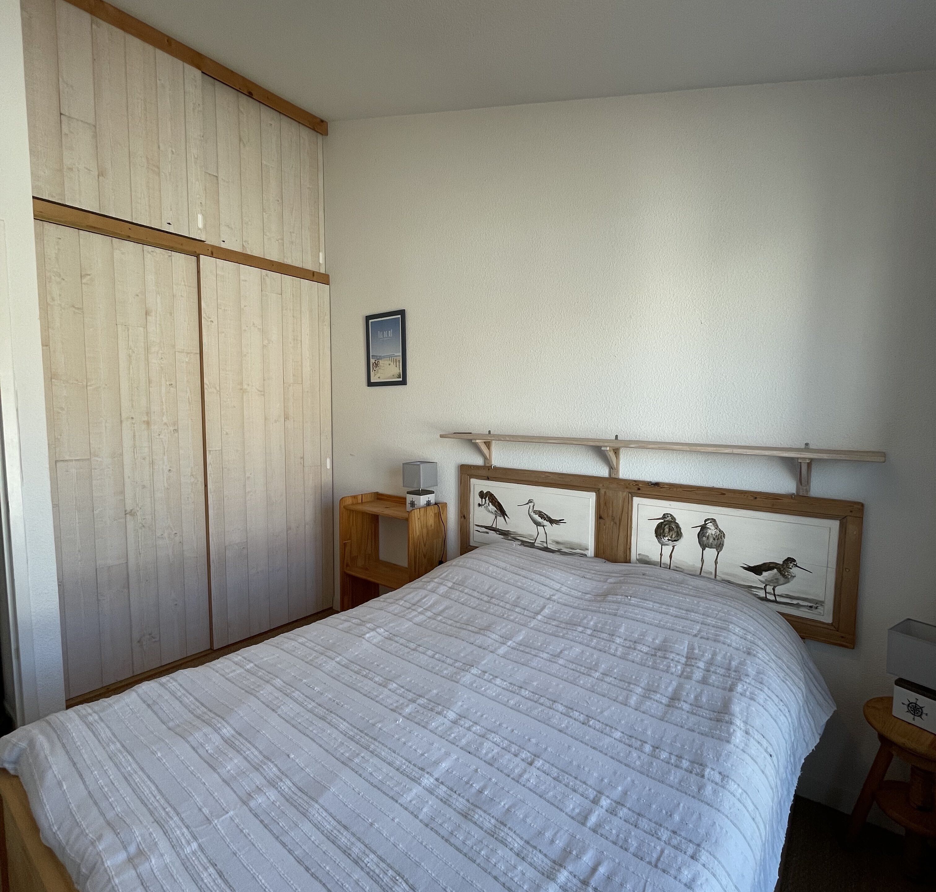A bedroom with a bed, wooden wardrobe, framed artwork, and a nightstand.