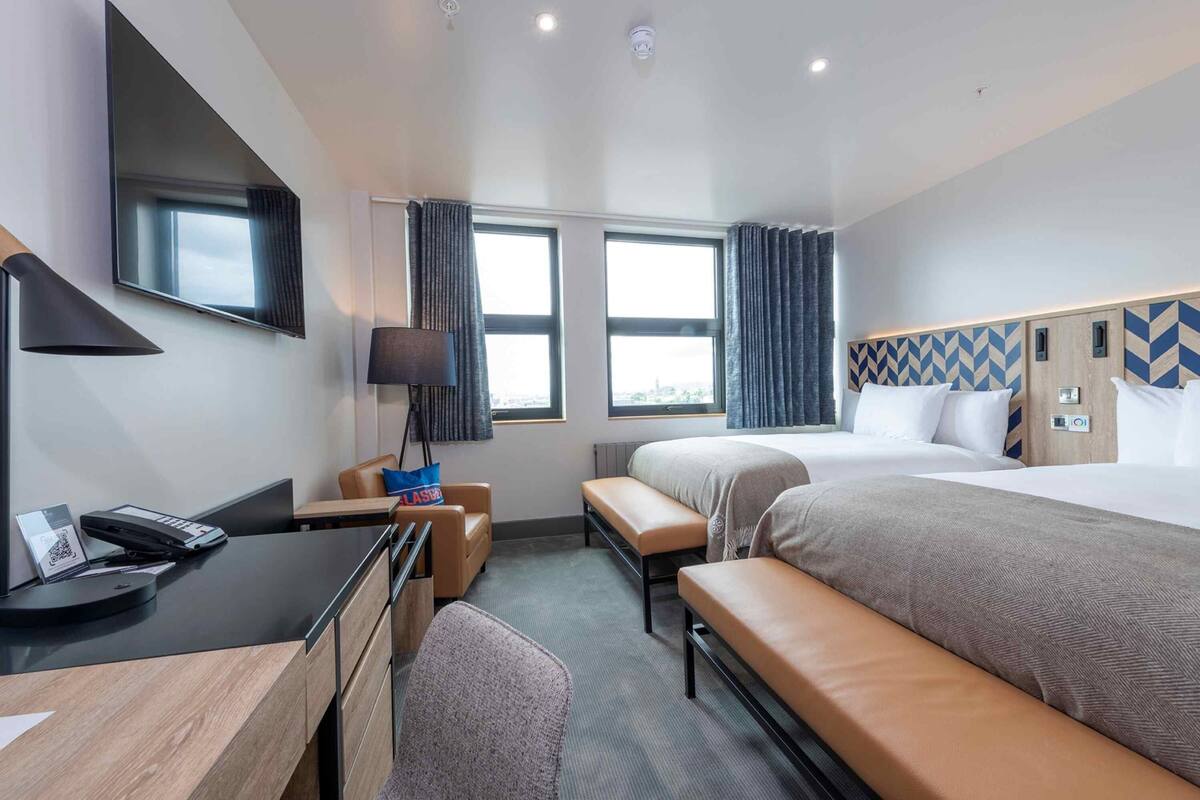 A hotel room with a desk, two beds, a TV, and a window with curtains.