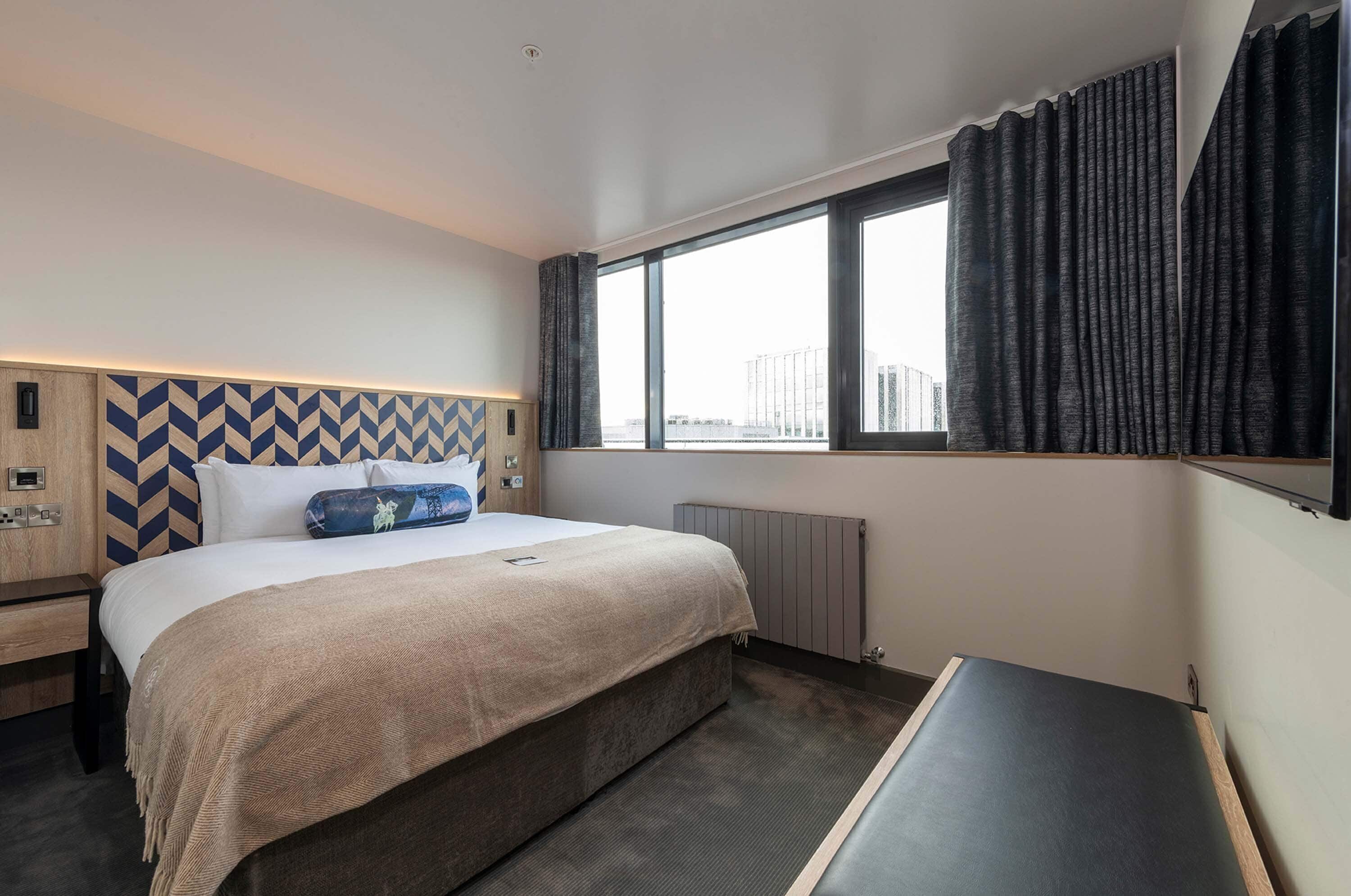 Ein Hotelzimmer mit Bett, Nachttisch, einem Fenster mit Vorhängen und Blick auf die Stadt.