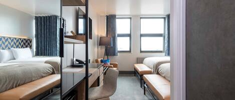 A hotel room with two beds, a desk with a chair, a lamp, and a view of the city through large windows.