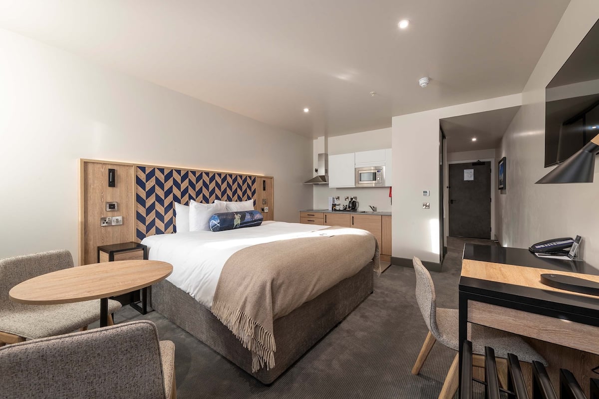 A modern hotel room with a large bed, a desk, and a kitchenette.