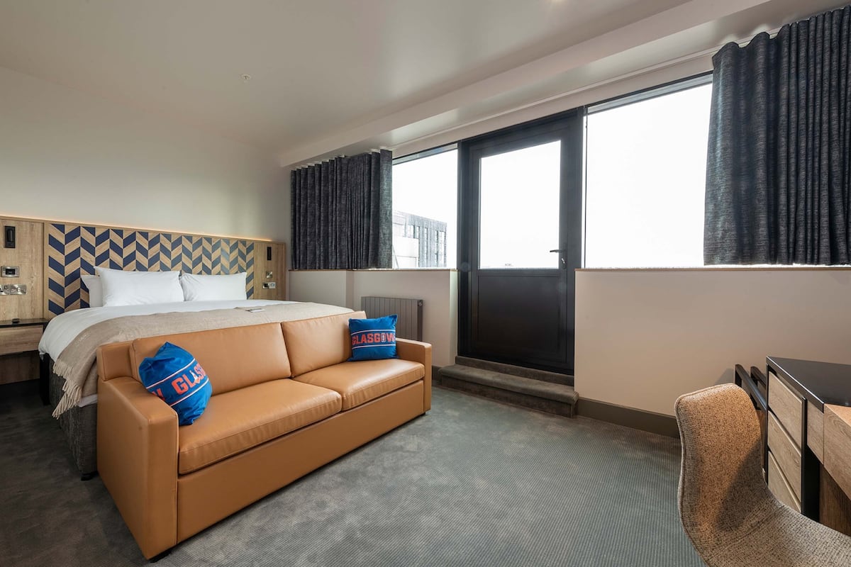 A hotel room with a bed, a brown leather sofa, a desk, and a large window with curtains.