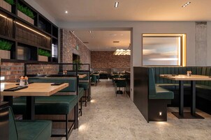 Un interior de restaurante moderno con mesas de madera y cabinas tapizadas de verde, paredes de ladrillo y una escalera visible.