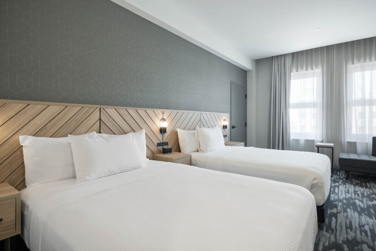 A hotel room with two beds, a wooden headboard, bedside lamps, a bench, and a large window with curtains.