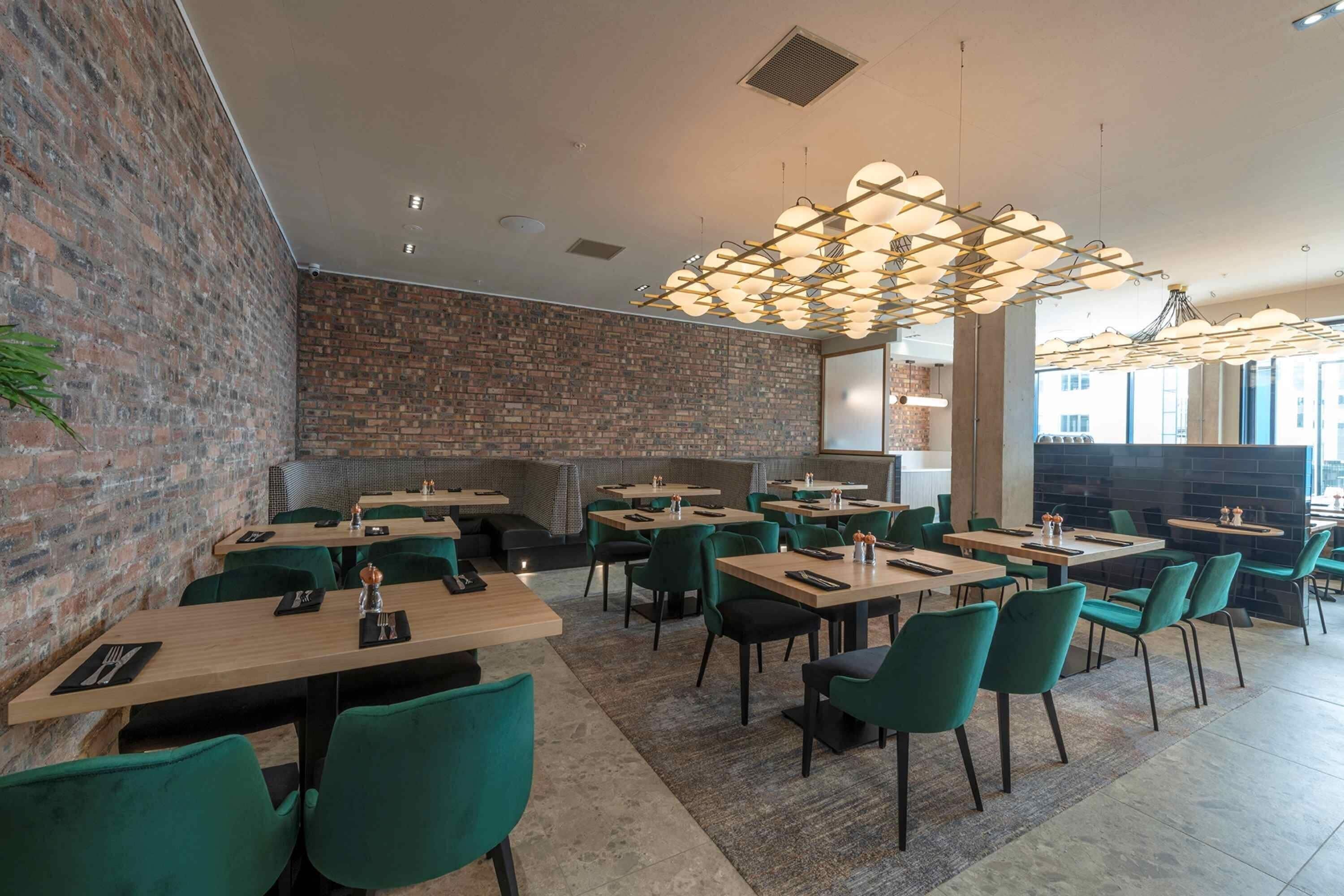 A restaurant with brick walls, green chairs, and modern pendant lighting. Tables are set with cutlery and napkins.