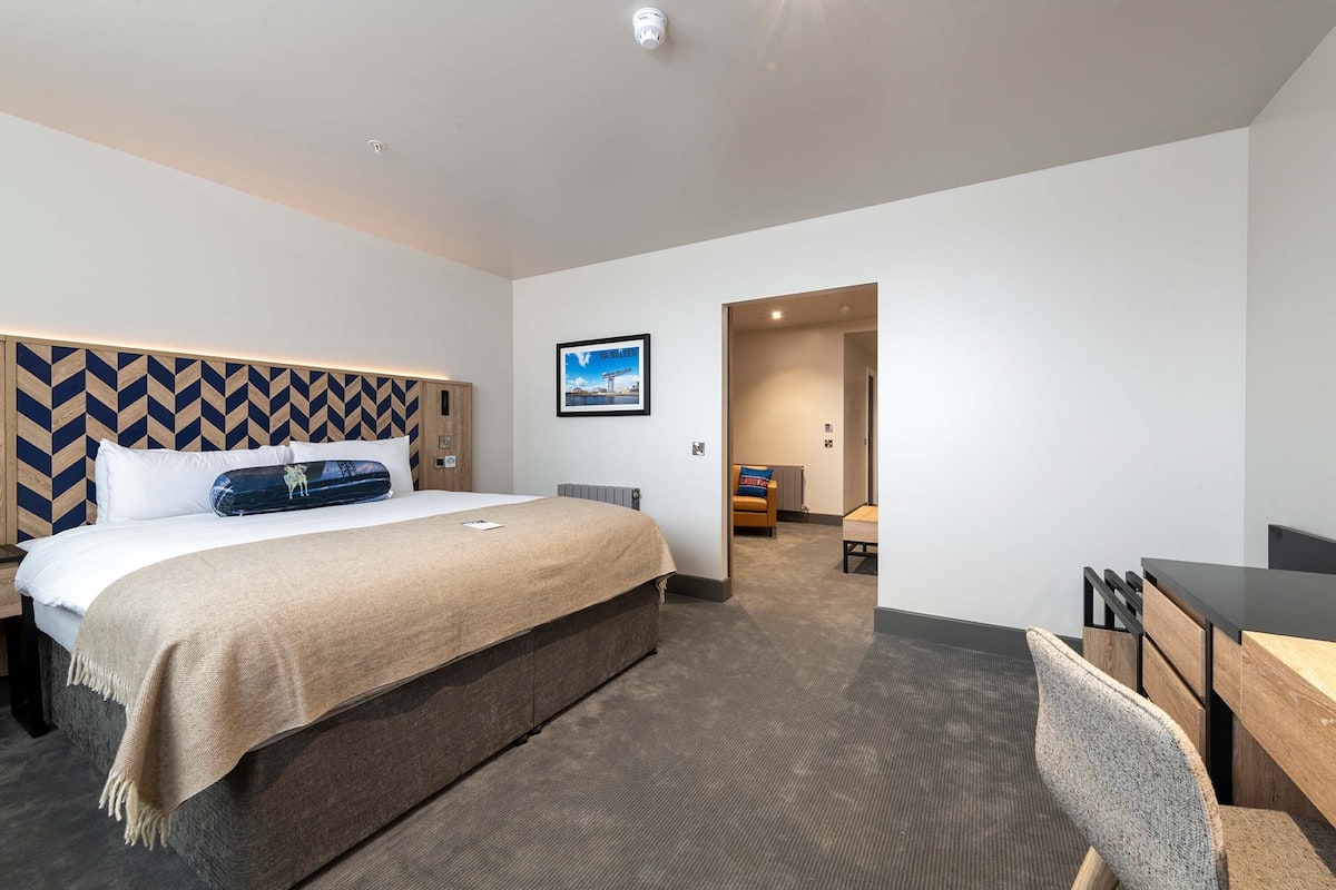 A hotel room with a large bed, a desk, and a chair. There is a picture on the wall and a door leading to another room.