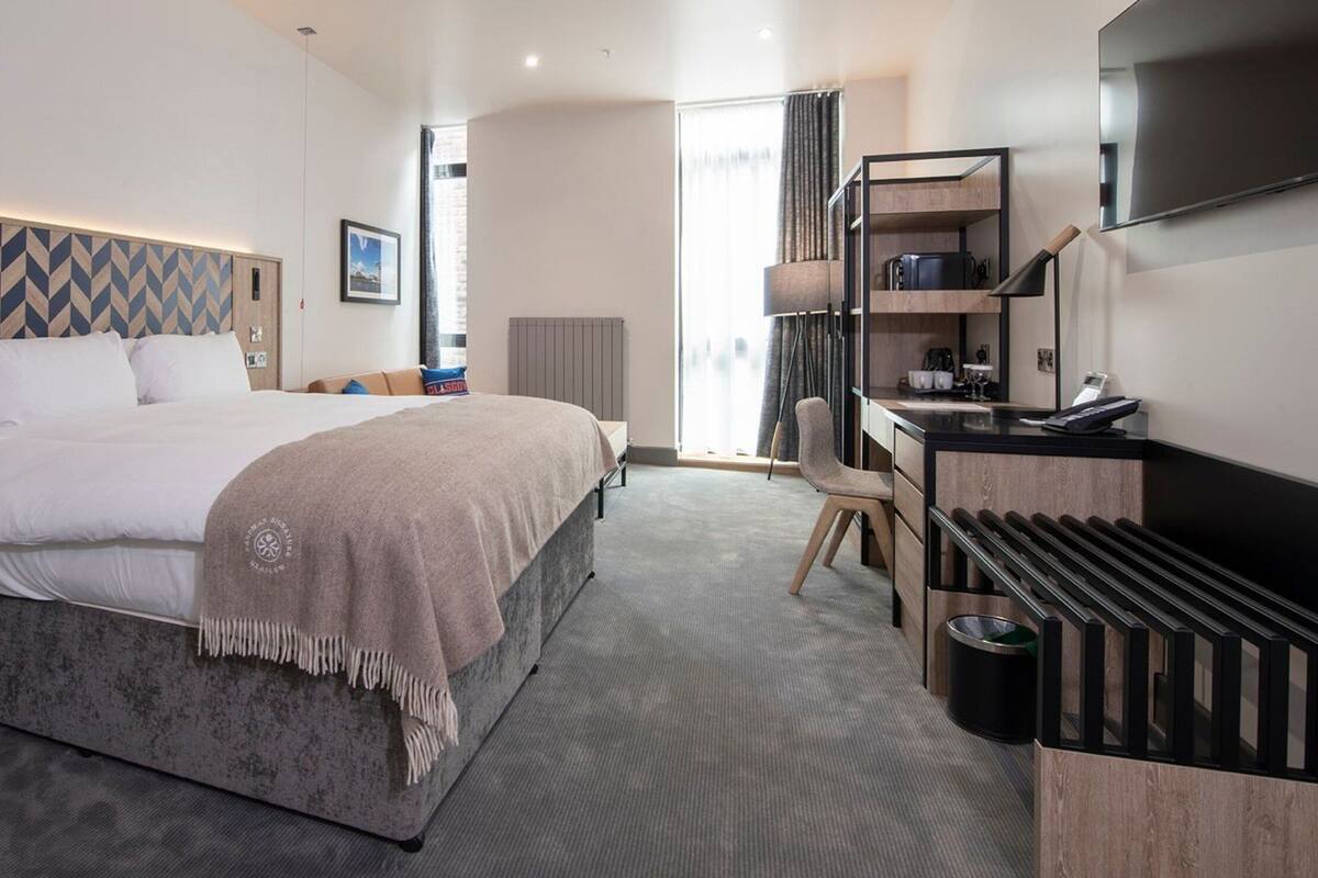 A modern hotel room with a large bed, a desk, a TV, and a minibar.
