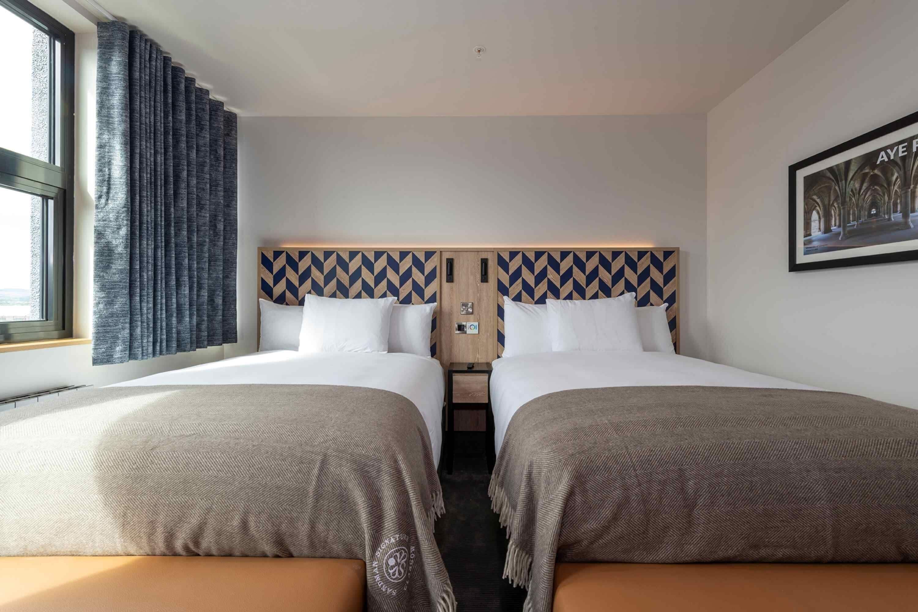 Une chambre d’hôtel avec deux lits, une grande fenêtre avec des rideaux, une tête de lit à motifs géométriques, un tableau encadré au mur et une petite table entre les lits.