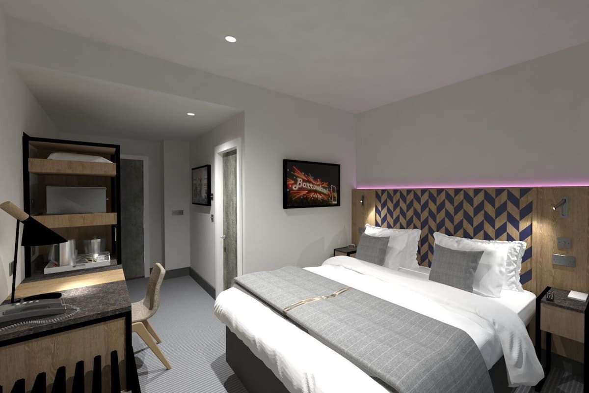 A modern hotel room with a bed, bedside tables, a desk with a chair, a wall-mounted TV, and a door leading to another room.