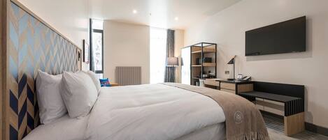 A modern hotel room with a large bed, a desk, a chair, a TV mounted on the wall, and a window with curtains.
