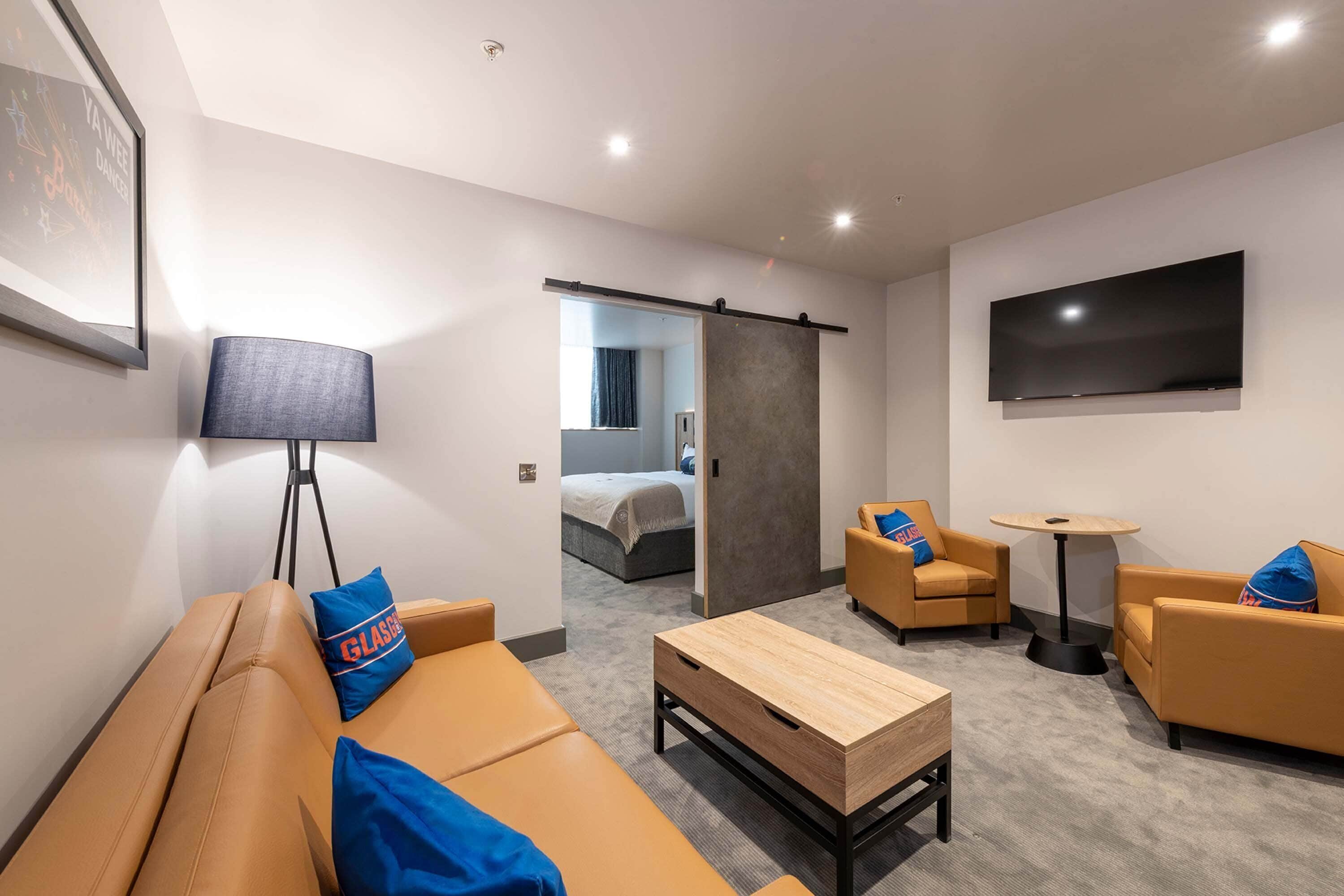 Une chambre d’hôtel moderne équipée d’un canapé, de fauteuils, d’une table basse, d’un téléviseur et d’une porte coulissante donnant sur une chambre à coucher.