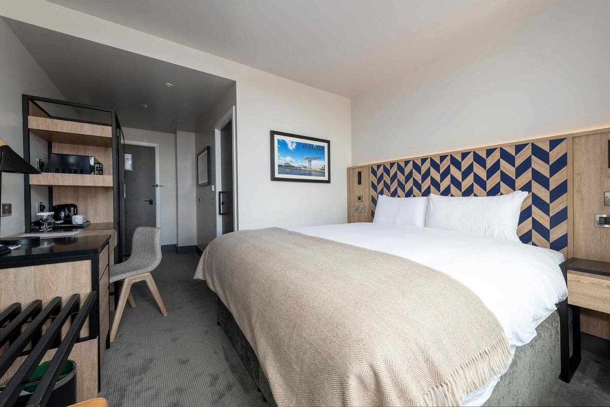 A hotel room with a bed, a desk, a chair, and a kitchenette.