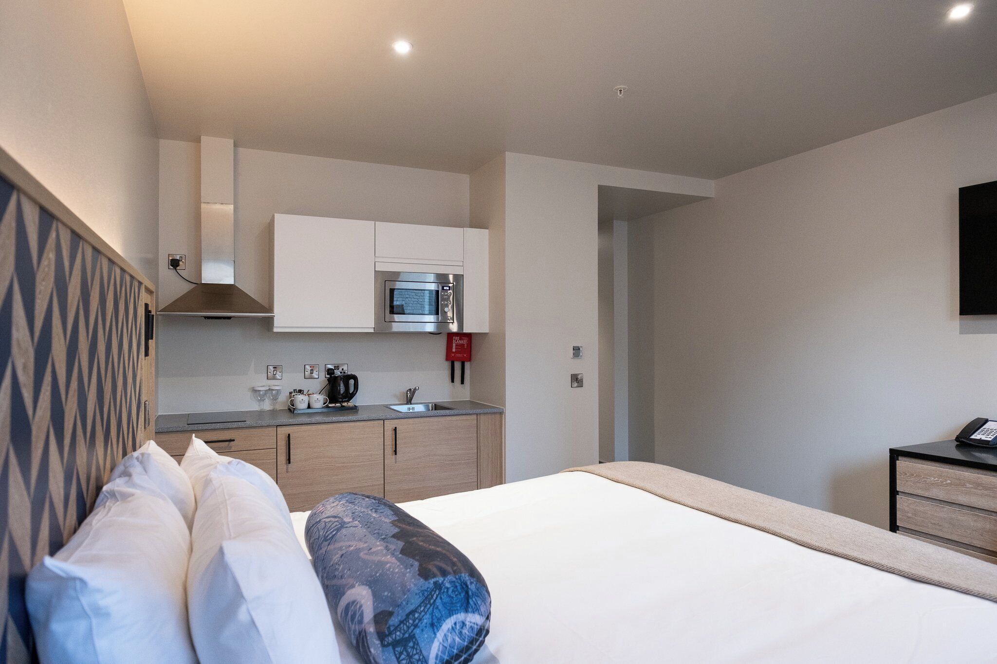 A modern hotel room with a bed, a small kitchenette, and a desk with a telephone.