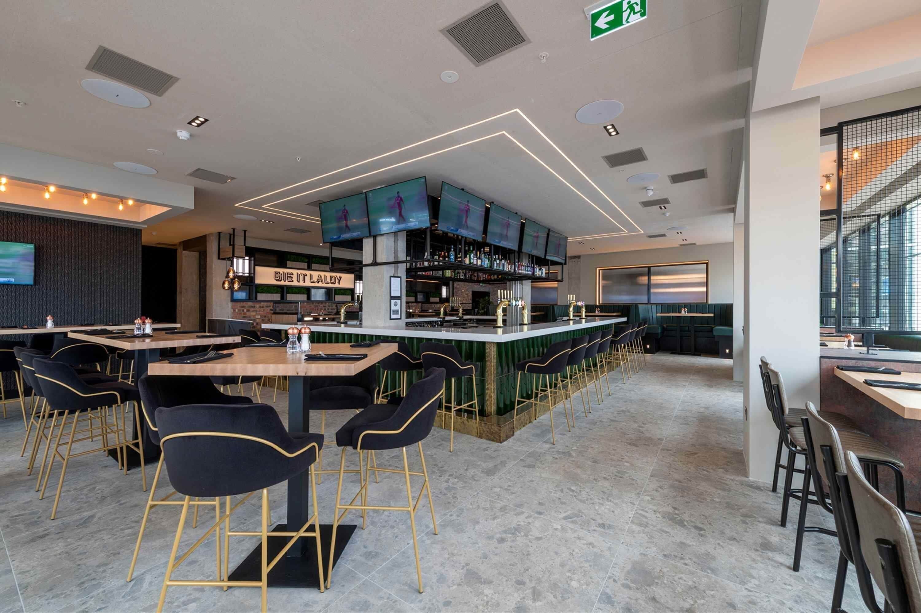 Un espace bar moderne doté de plusieurs écrans, d’un comptoir et de différents types de sièges.