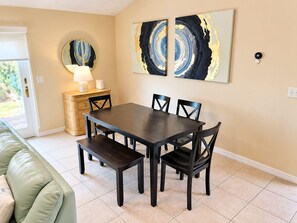 A dining area with a dark wooden table and chairs, a cream-colored wall, and abstract art pieces. - Bikini Bungalow - King Beds and Short Walk to the Beach! Pet Friendly! (St. Augustine)