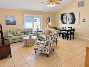 A living room with a sofa, armchair, coffee table, TV stand, dining table, chairs, artwork, and a ceiling fan. - Bikini Bungalow - King Beds and Short Walk to the Beach! Pet Friendly! (St. Augustine)