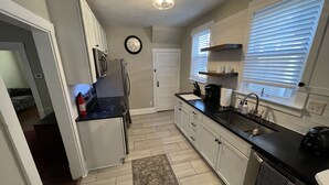 A modern kitchen with white cabinets, a black countertop, a stainless steel refrigerator, a microwave, and a built-in oven. There is a window with blinds, a clock on the wall, and a rug on the floor. - Newly Updated: Nearby Short North / OSU! (Columbus)