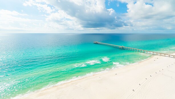 Ein Strand mit einer langen Pier, die sich in das türkisblaue Meer erstreckt, unter einem teils bewölkten Himmel.