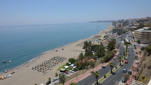 Paisagem costeira com praia, carros estacionados e edifícios à beira-mar.