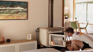 A cozy room with a wood-burning stove, a painting of a beach scene, and a view of the outdoors through a large window.