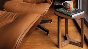 A tan leather sofa and armchair in a cozy room with a wooden floor, a small wooden side table with a book and a mug, and a metal shelving unit against the wall.