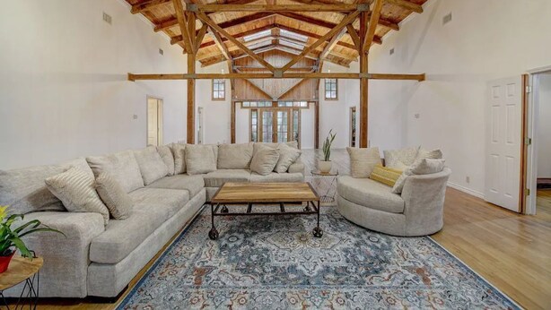A spacious living room with a large L-shaped sectional sofa, a wooden coffee table, and a patterned rug. The room features exposed wooden beams on the ceiling and large windows allowing natural light.