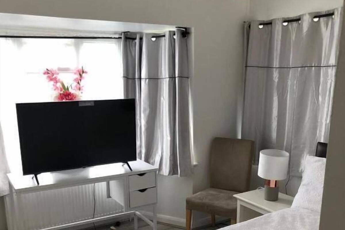A hotel room with a bed, a desk with a TV, a chair, a nightstand with a lamp, and curtains.