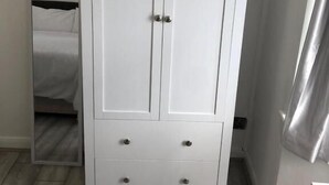 A white wardrobe with two doors and a wooden top shelf, located in a room with a bed and a curtain. - Featherstone Court (London)