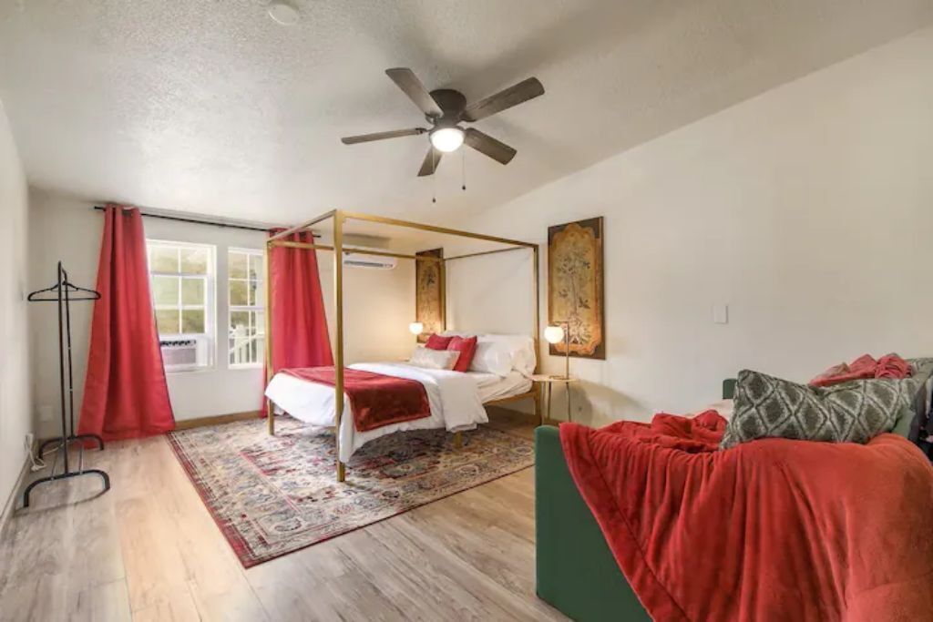 A four-poster bed with a canopy, a bedside table with a lamp, a green sofa, and a red rug in a room with wooden flooring and a ceiling fan.