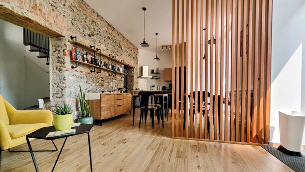 Een moderne woonkamer met een bakstenen muur, houten vloer en een gele bank. Er is een keuken met houten keukenkastjes en een eettafel.