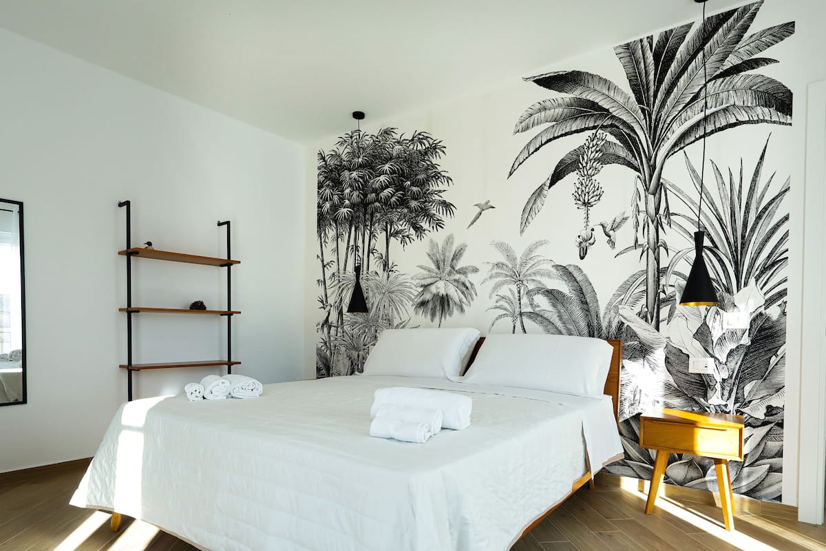 Een slaapkamer met een bed, een houten nachtkastje, een zwarte hanglamp en een muurschildering van tropische planten.
