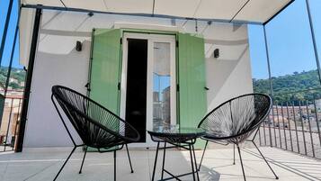 A balcony with two black metal chairs and a table, a green door, and a view of buildings and hills in the background.