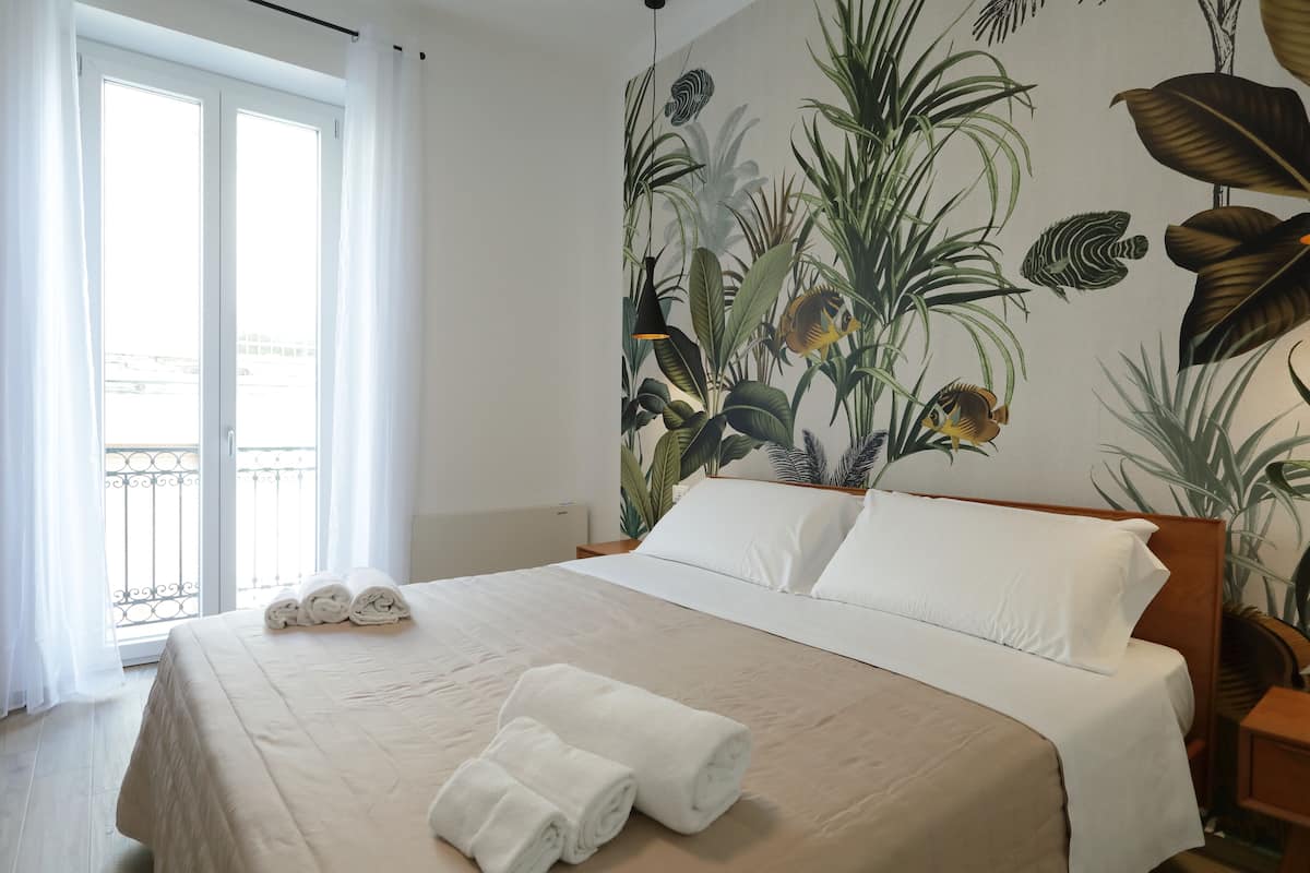 Een hotelkamer met een bed, witte lakens, een tropische muurschildering en een balkon met witte gordijnen.