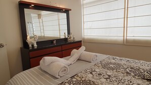 A neatly made bed with rolled towels, a large mirror above a dark wooden dresser, and a window with blinds.