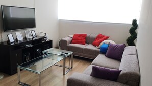 A modern living room with a flat-screen TV, a glass coffee table, a grey sofa with colorful cushions, and a black entertainment unit with framed photos.