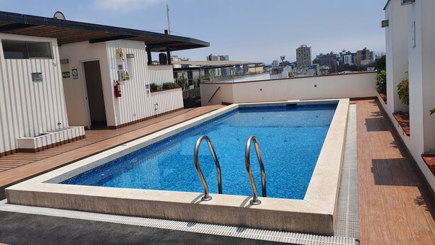 A rooftop swimming pool with metal ladders, surrounded by a tiled deck and a white building with a striped facade.