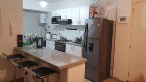A compact kitchen with white cabinets, a stainless steel refrigerator, a microwave, and a gas stove. There are three black bar stools at the counter.