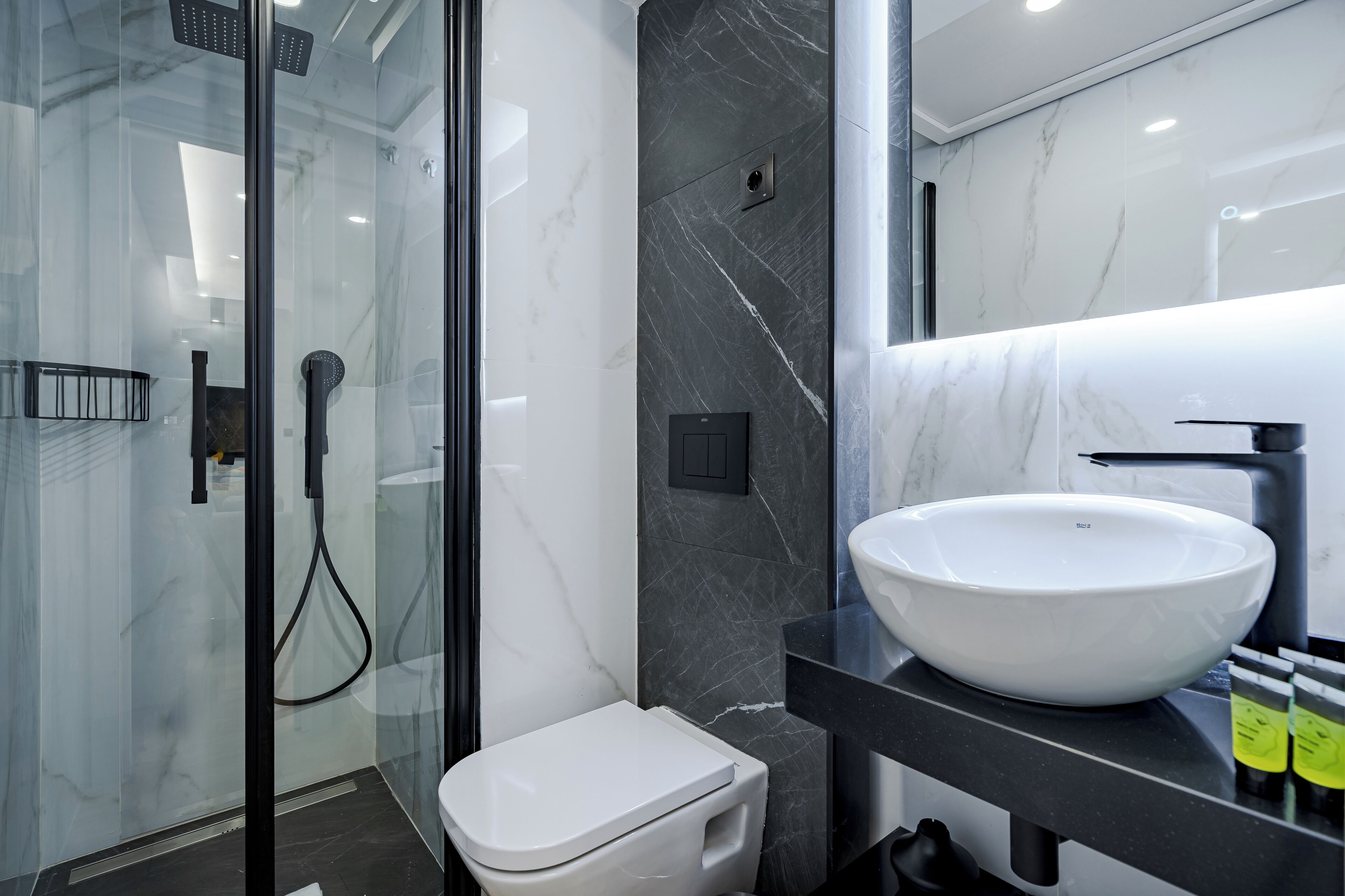 Une salle de bain moderne avec une douche à parois de verre, des toilettes blanches et un grand lavabo blanc posé sur un plan de travail noir.