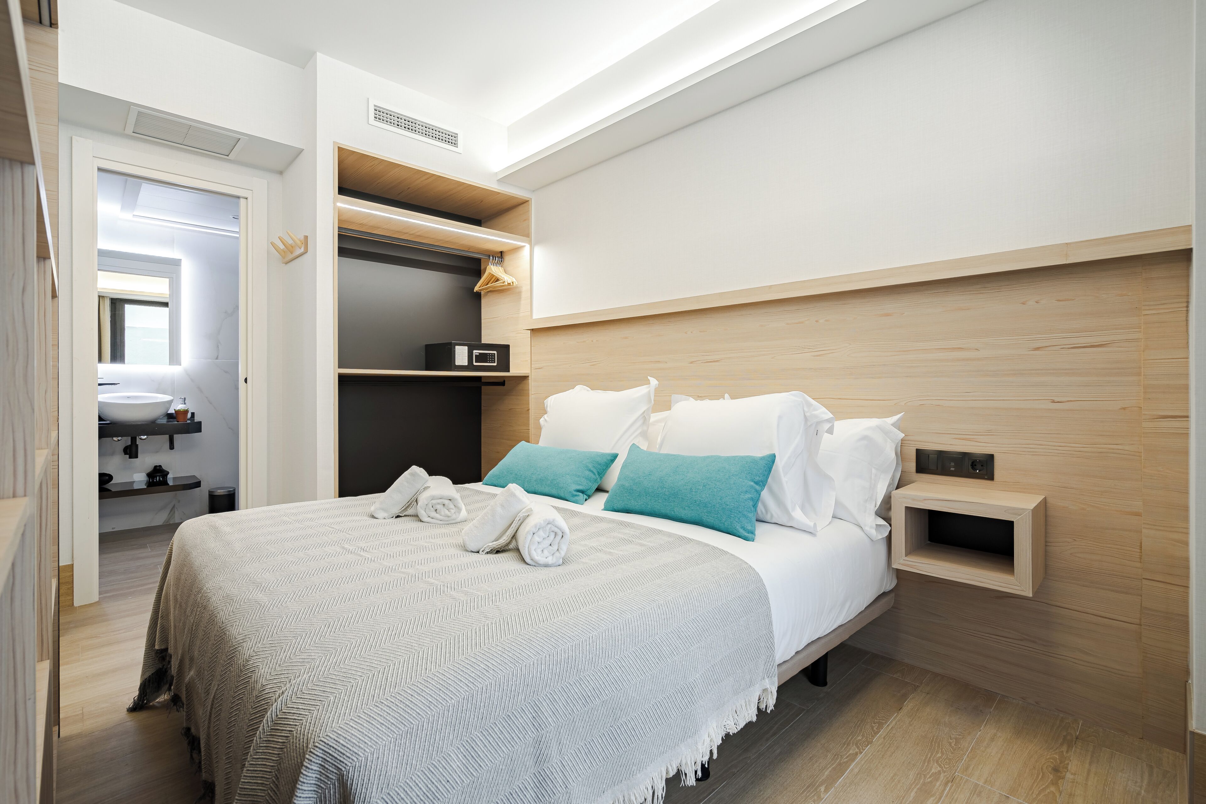 A modern hotel room with a large bed, a built-in wardrobe, a bedside table, and a small shelf.