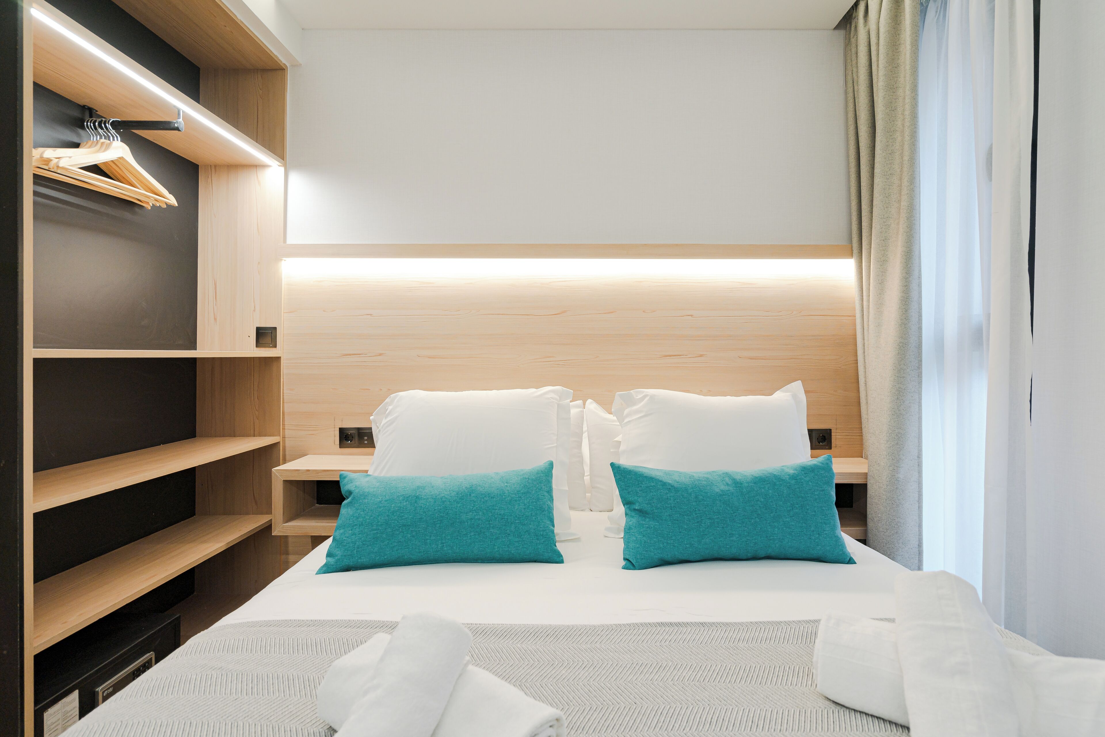 Una habitación de hotel con una cama, dos almohadas, una mesita de noche, un estante de madera con perchas y una ventana con cortinas.