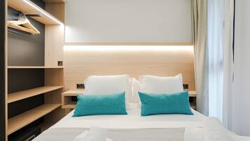 Una habitación de hotel con una cama, dos almohadas, una mesita de noche, un estante de madera con perchas y una ventana con cortinas.