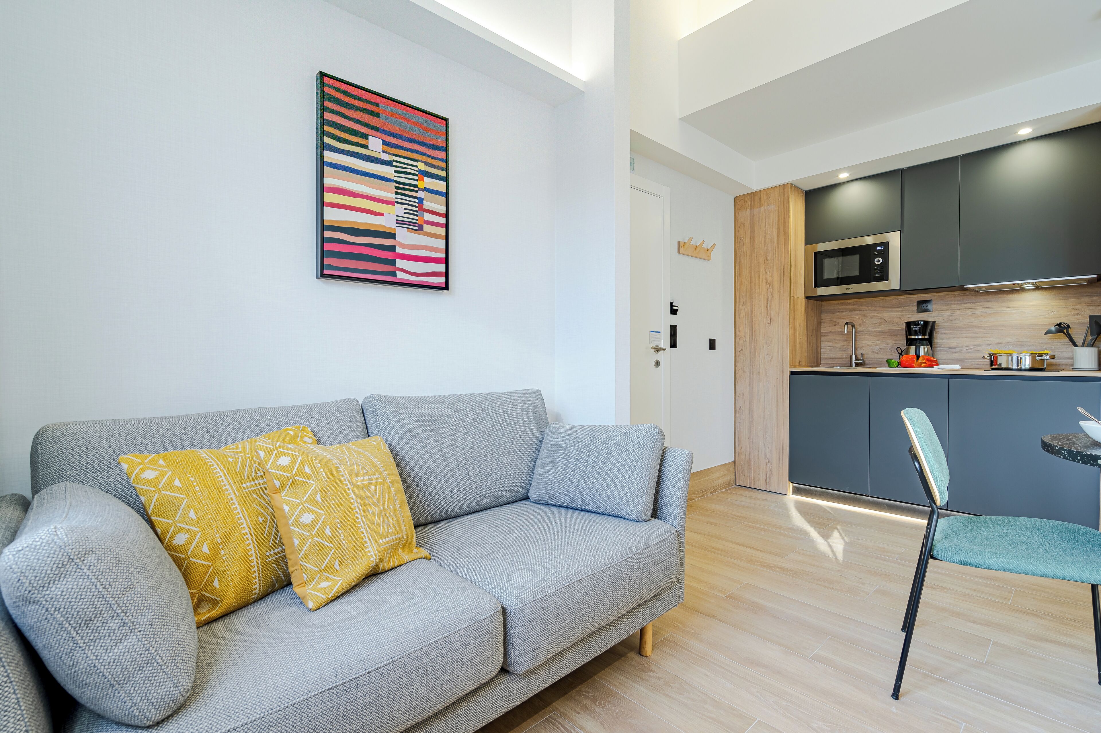 A modern living room with a grey sofa, a colorful abstract painting, and a kitchen area with dark cabinets and a microwave.