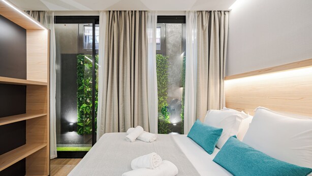 A modern hotel room with a bed, a wooden shelf, and a view of greenery through a glass door. - Feelathome Ventas Suites (Madrid)