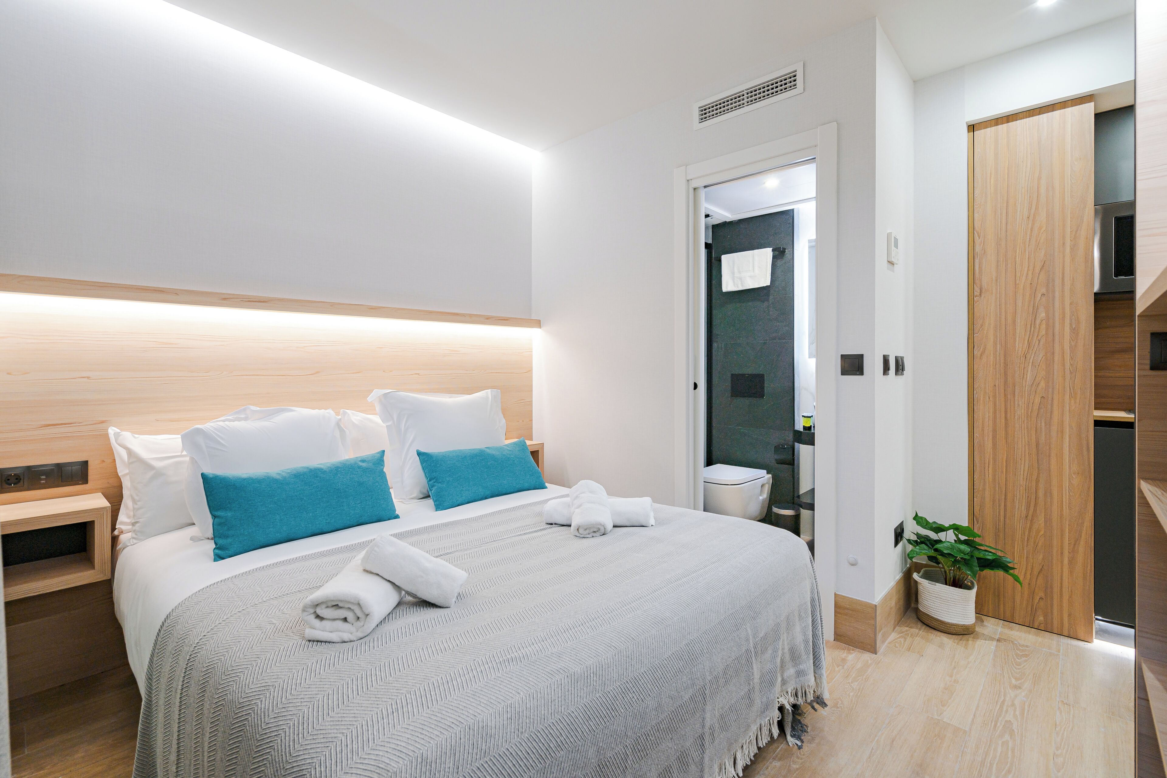 A modern hotel room with a large bed, a bedside table, a potted plant, and a wooden wardrobe.