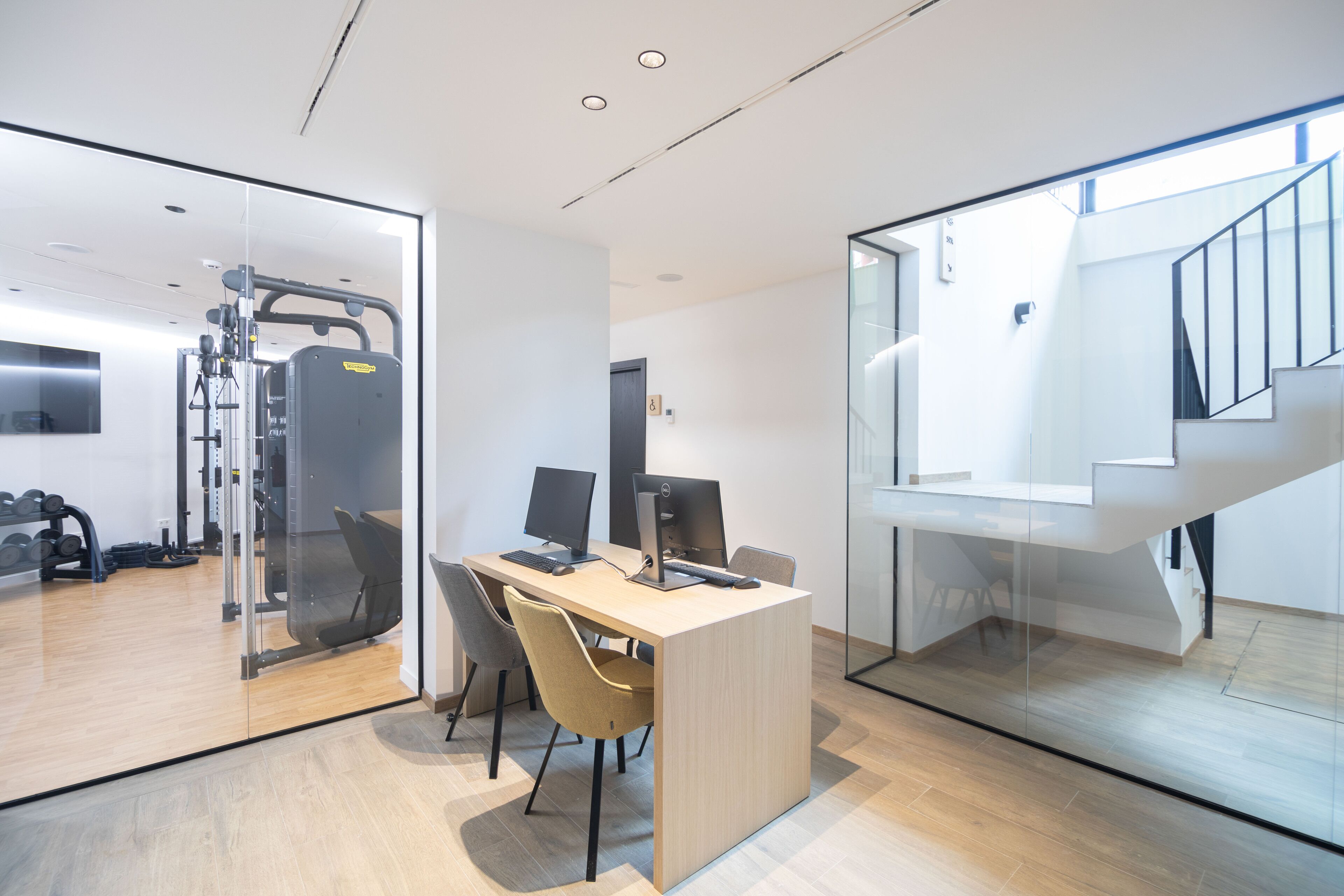 A modern office space with a desk, computers, and a gym area visible through glass partitions.