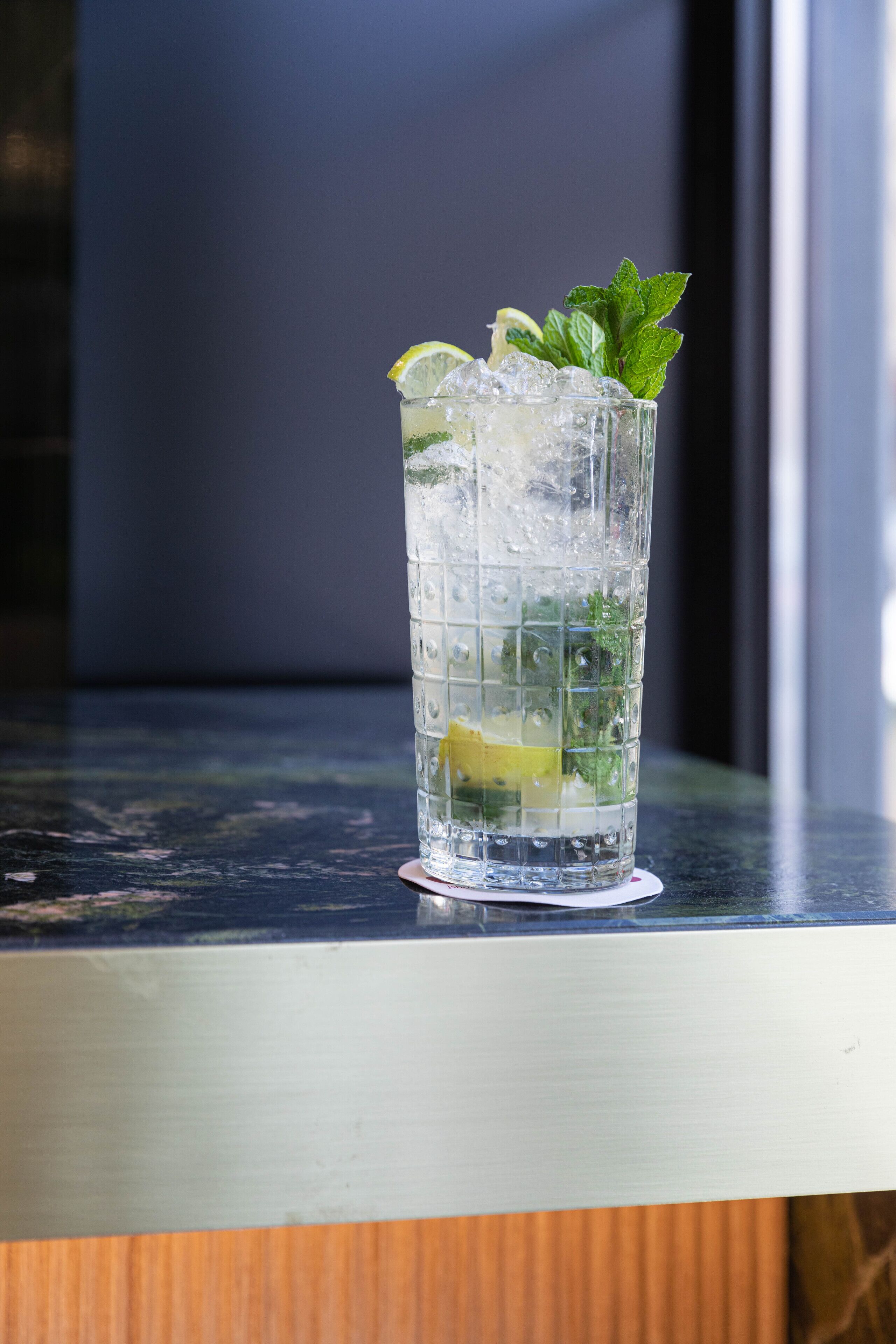 A tall glass with ice cubes, lemon slices, and mint leaves on a table.