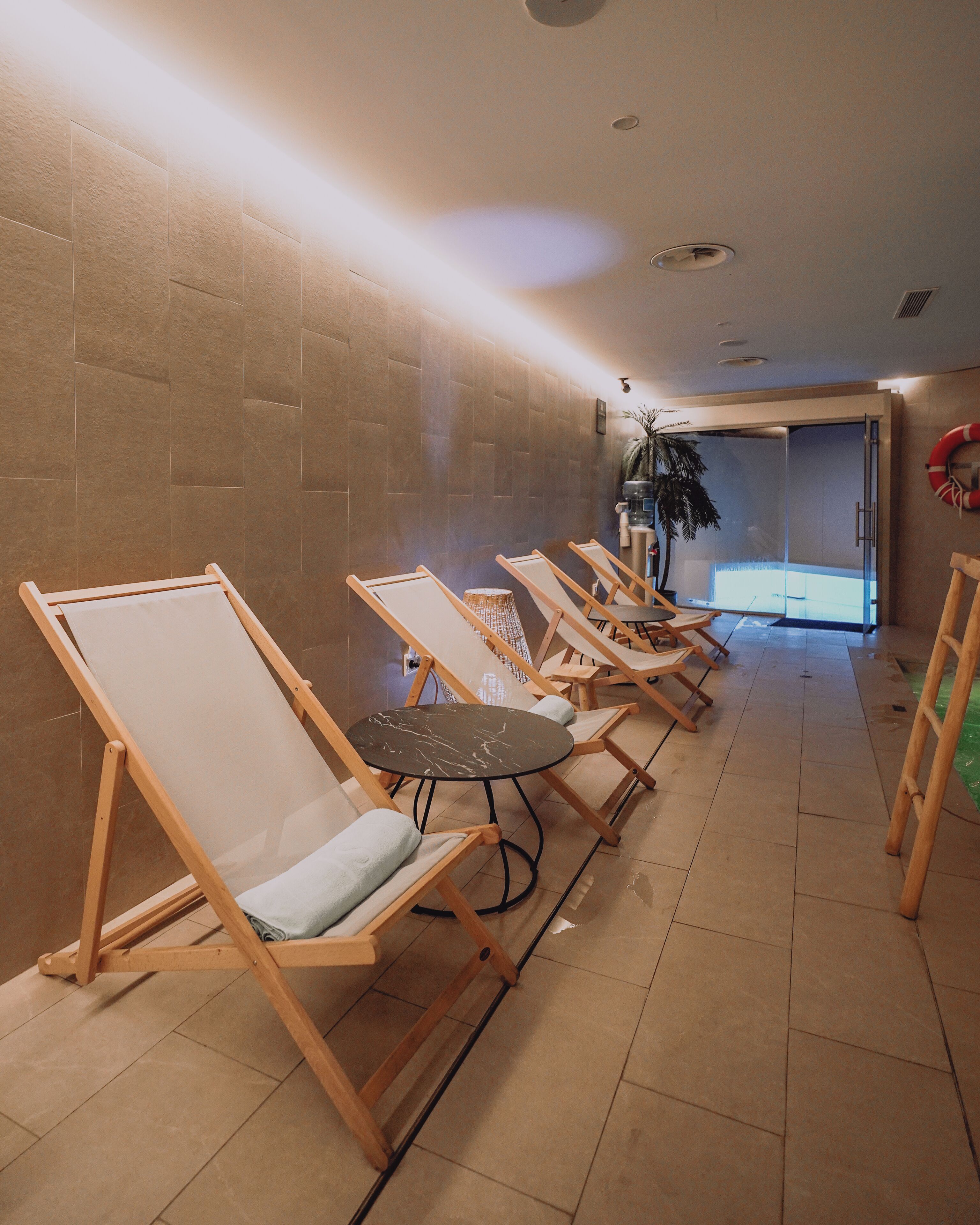 A room with multiple wooden lounge chairs arranged in a row, a small round table, and a wall-mounted plant.