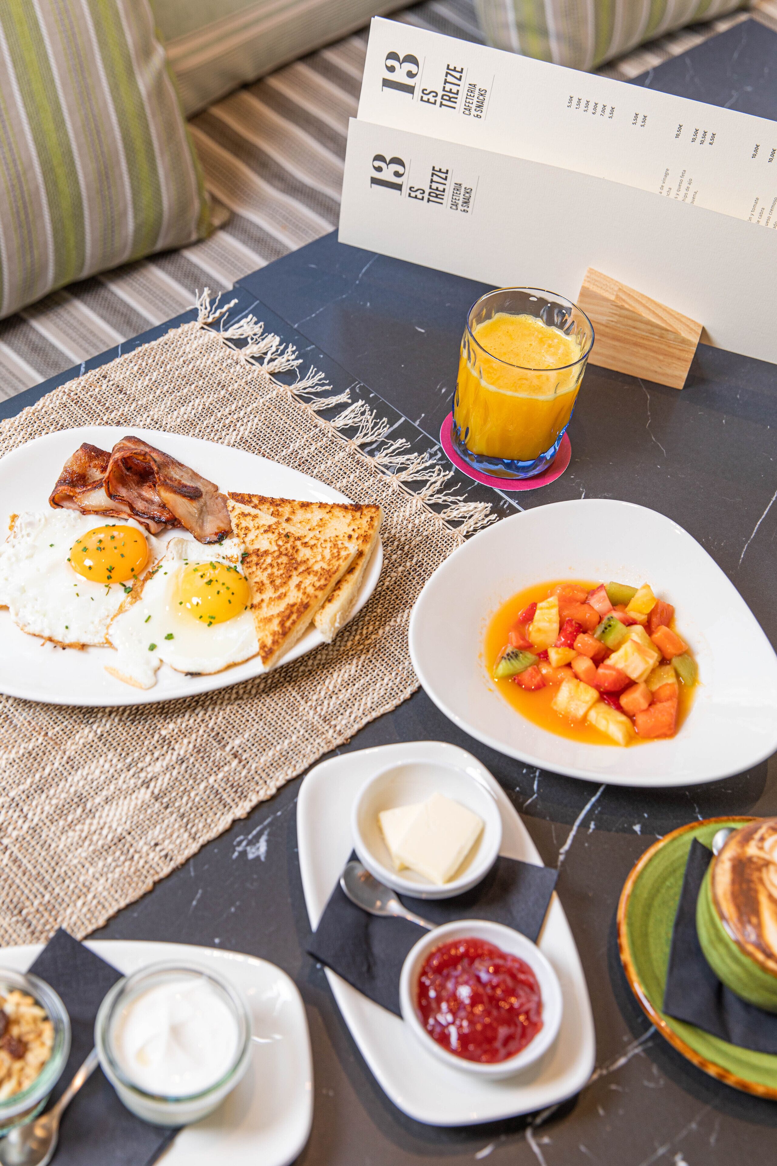 Desayuno con tostadas, huevos, tocino, un vaso de jugo de naranja y un tazón de ensalada de frutas sobre una mesa.