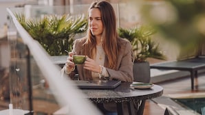 Una persona sentada a una mesa con una computadora portátil y una taza verde, con vistas a un paisaje urbano con edificios y zonas verdes.